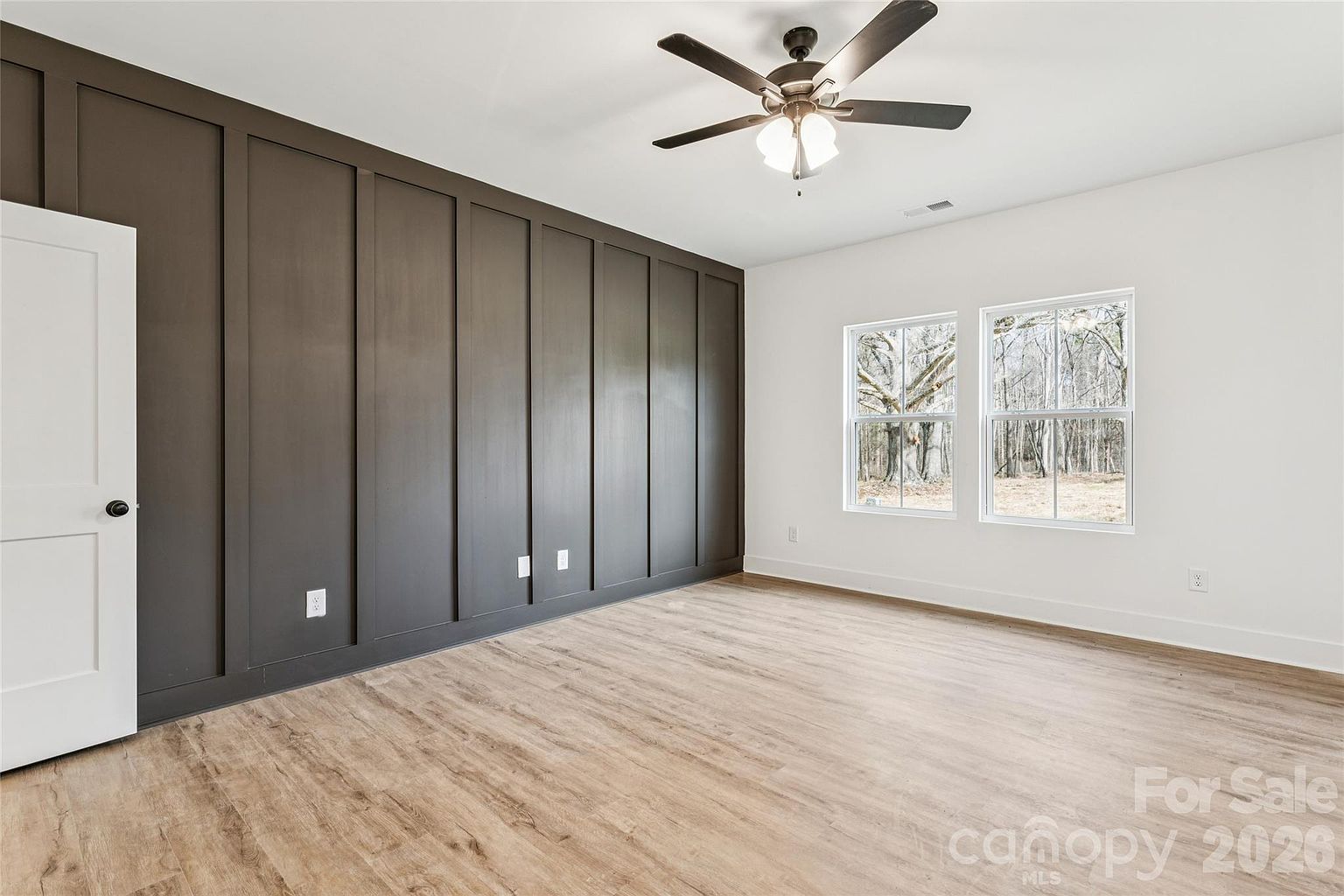 Primary bedroom with dark accent board-and-batten feature wall, ceiling fan, and windows facing the wooded backyard